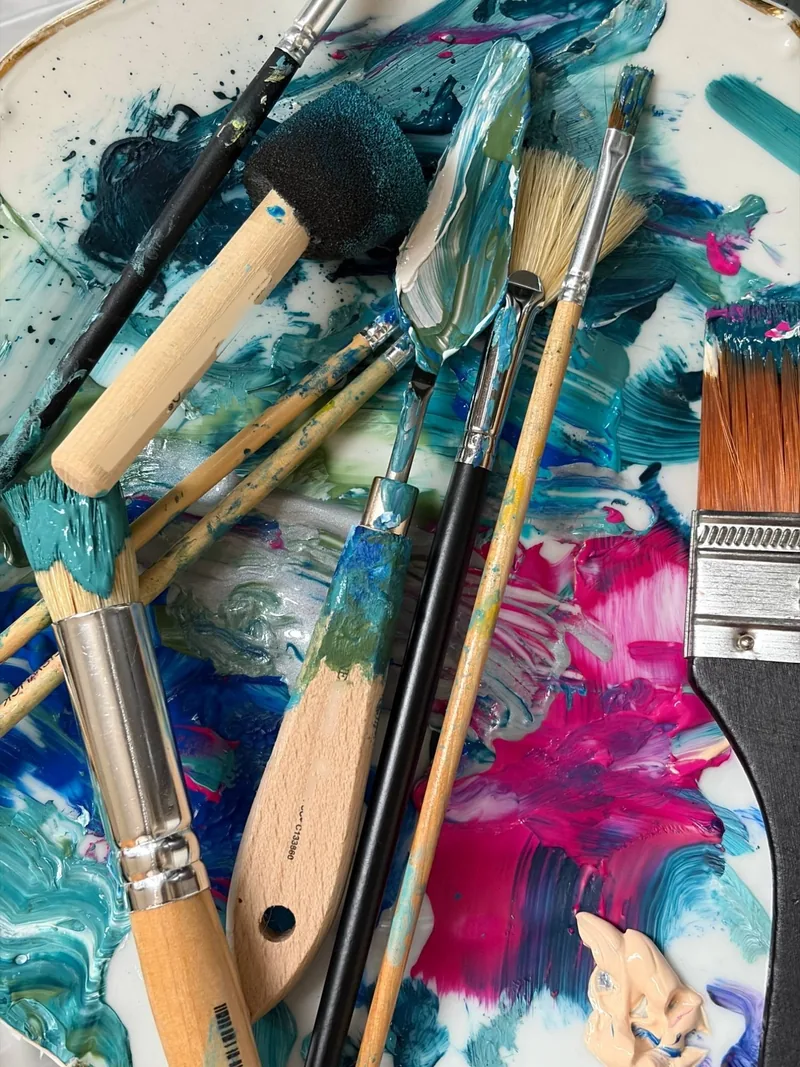 Brushes, a sponge and a palette knife on a palette full of turquoise, blue and pink paint